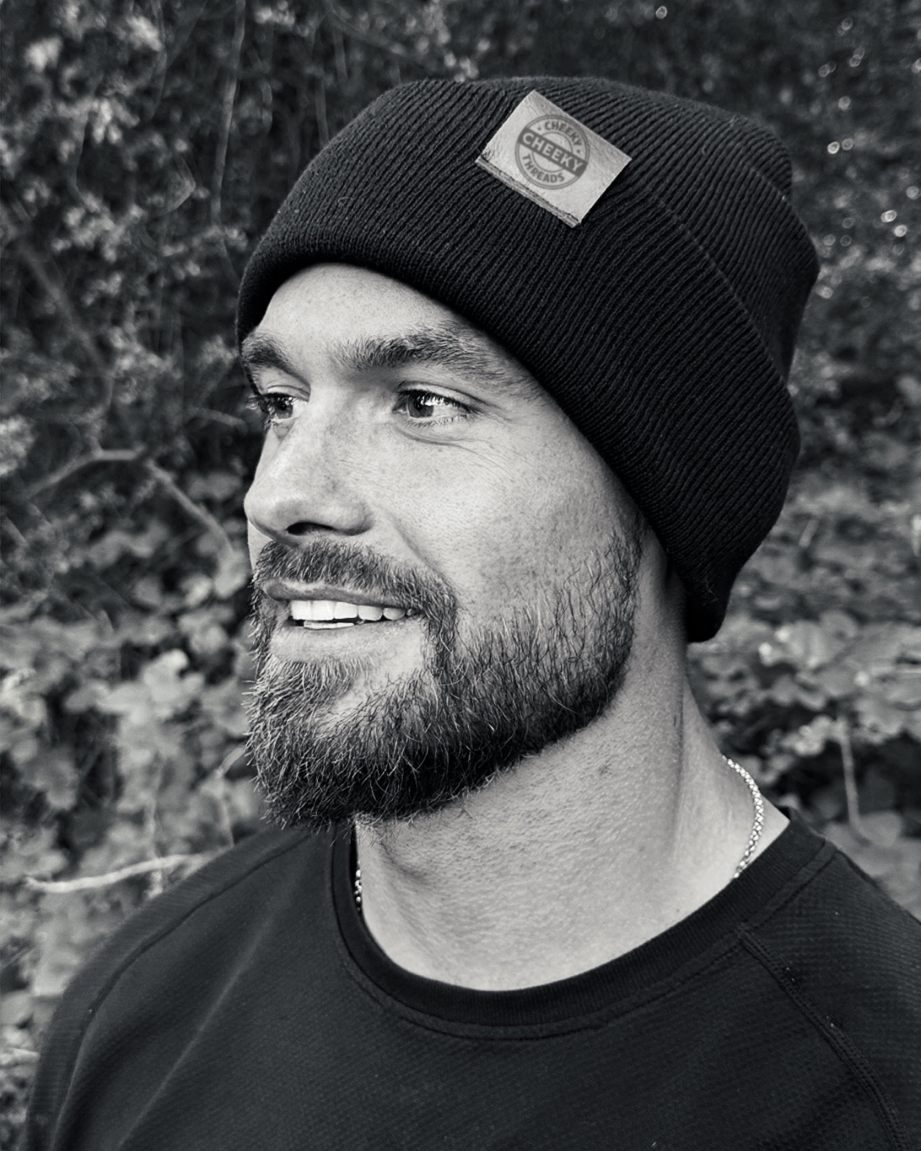 Man wearing a black beanie with a brand logo, standing against a natural background.