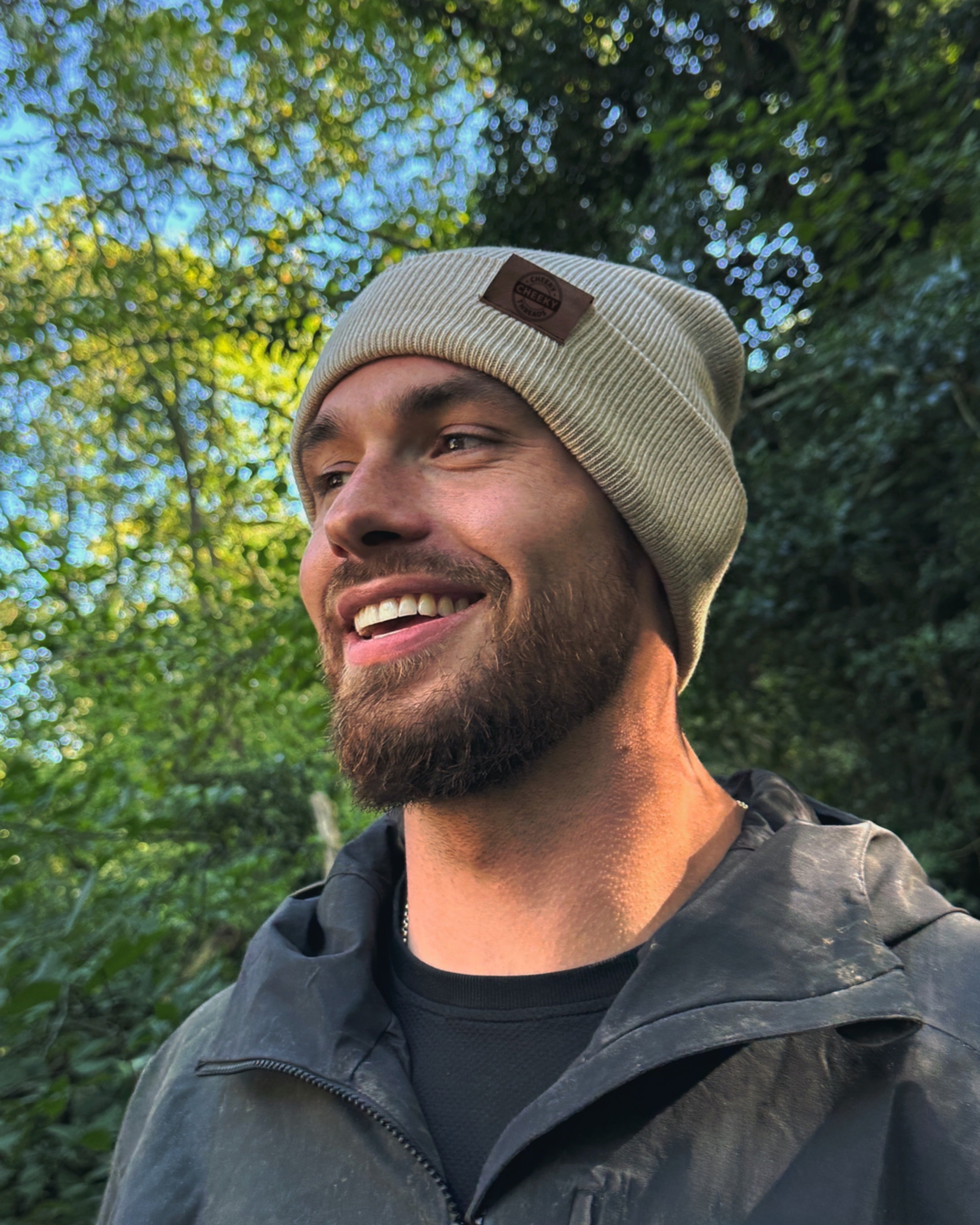 Man wearing a beige beanie with a logo, standing outdoors with trees in the background